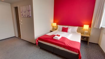 Una habitación de hotel con una cama grande, dos lámparas de noche, una pared de acento roja y una puerta que da a otra habitación.
