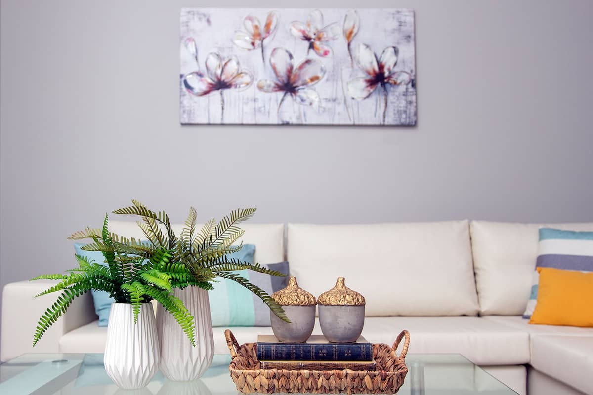 A living room with a white sofa, a glass coffee table, a fern plant, two decorative bowls, and a basket of books.