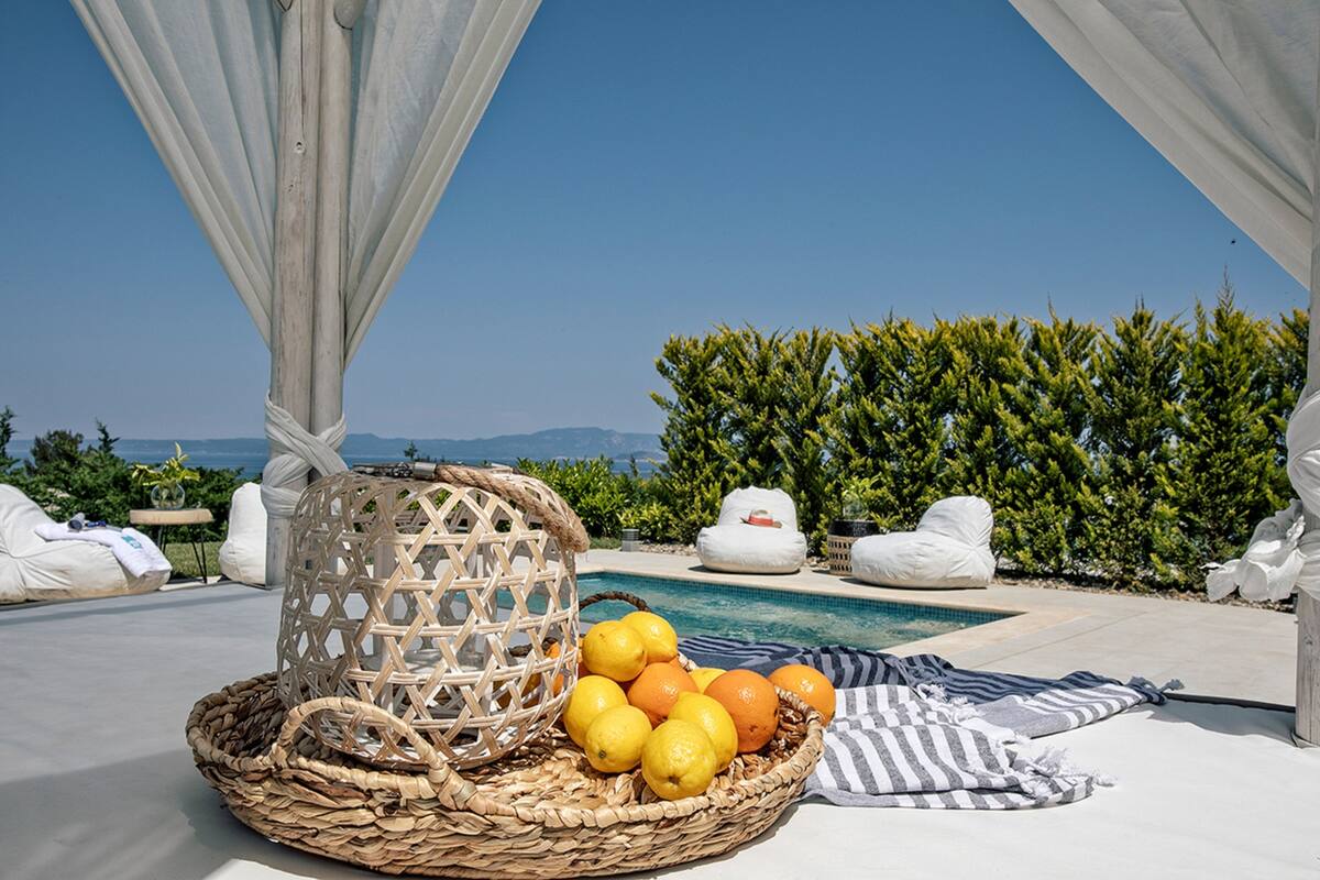A wicker basket with lemons and oranges on a poolside lounge area with white cushions and a view of the sea.