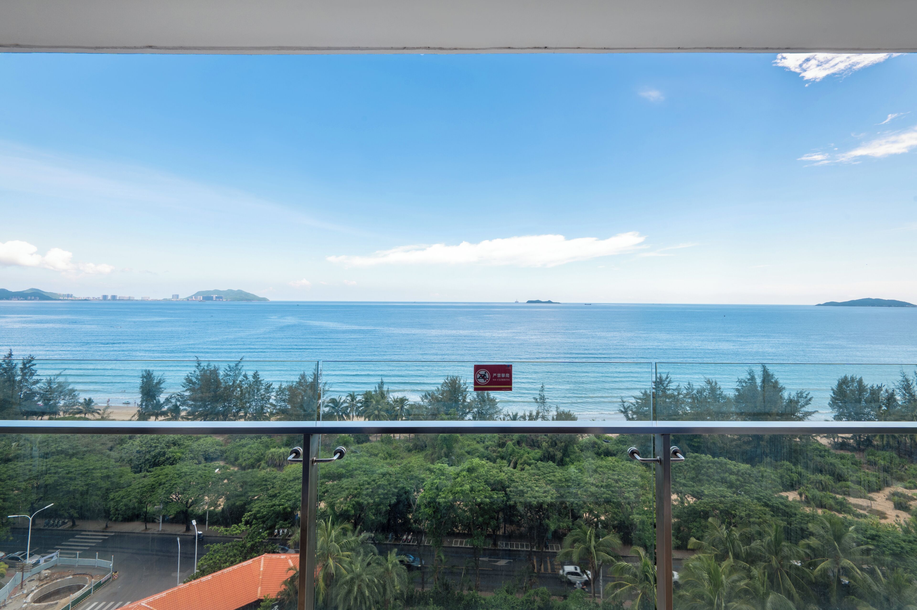 Une vue depuis un balcon donnant sur une plage, avec un ciel dégagé, un panneau indiquant qu'il est interdit de fumer et une végétation luxuriante.