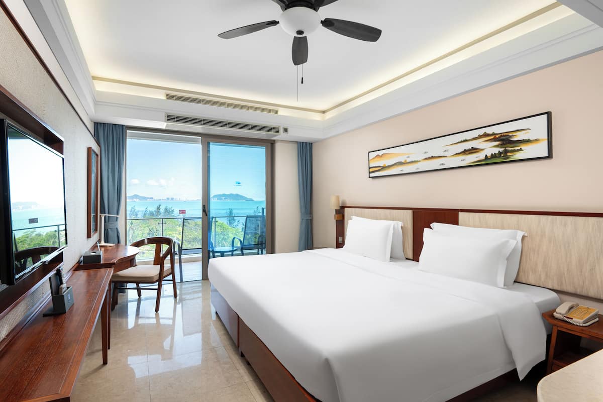 Una camera d'albergo con un letto grande, una scrivania con una sedia, una ventola a soffitto, un comodino con una lampada e un quadro appeso al muro.