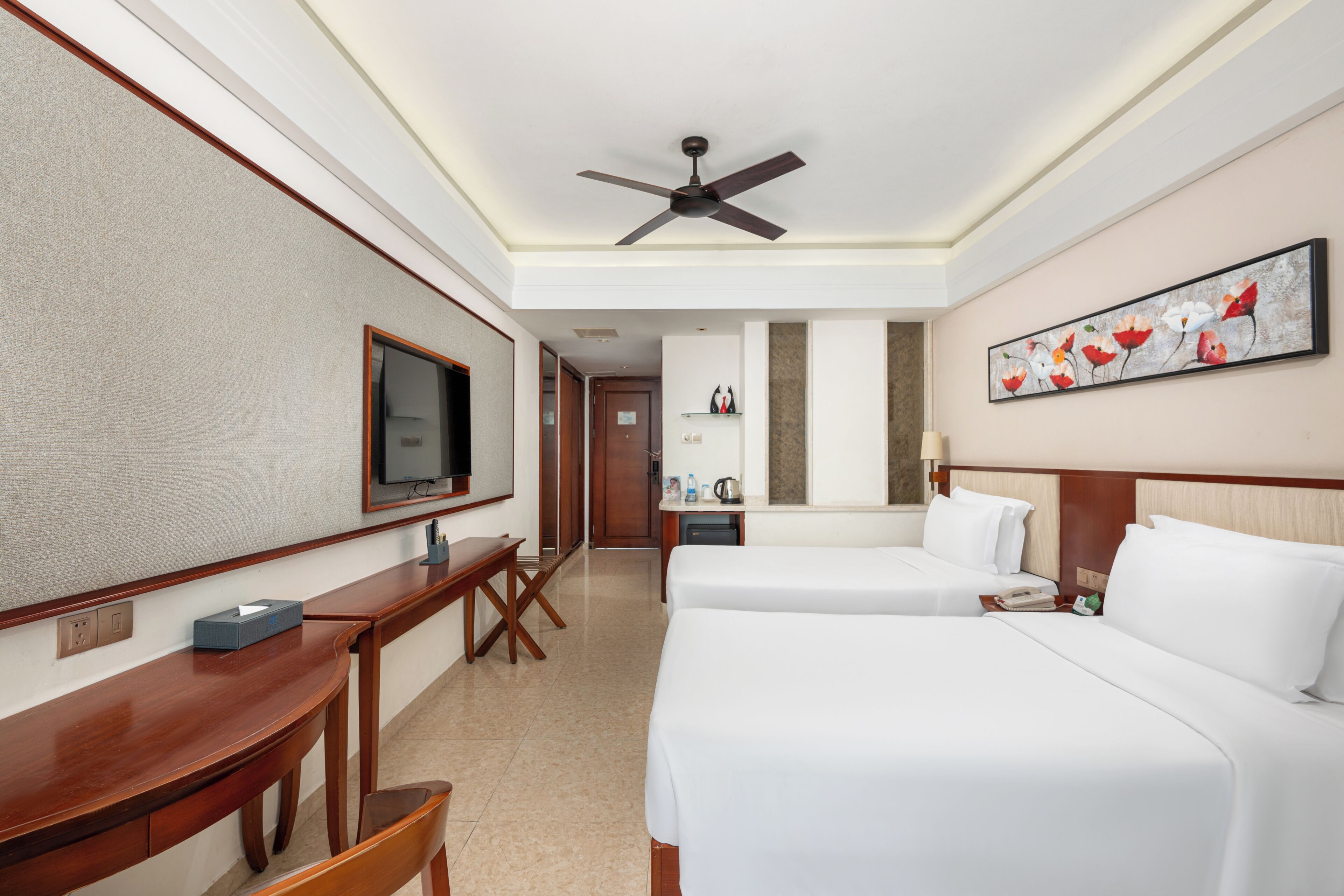 A hotel room with two beds, a desk, a chair, a television, and a ceiling fan.
