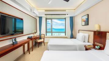 A hotel room with a bed, a desk, a chair, a television, a window with a view of the sea, and a ceiling fan.