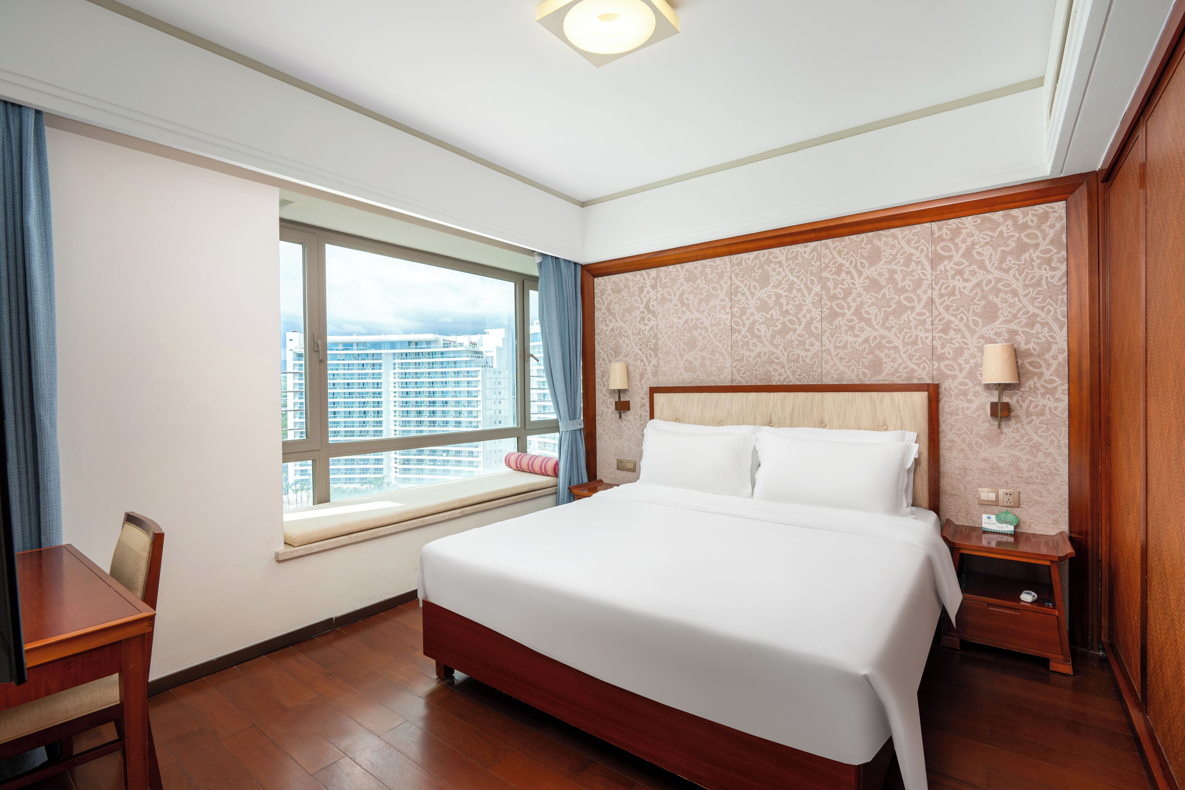 A hotel room with a large bed, a window with a view of buildings, a desk, and a nightstand with a lamp.