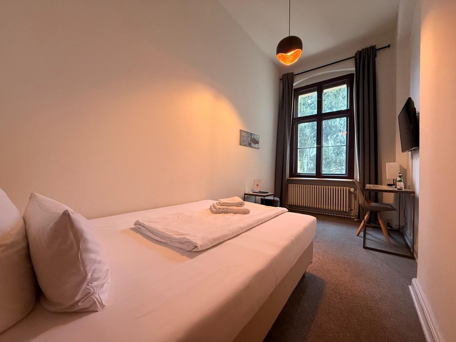 Ein Schlafzimmer mit Bett, Schreibtisch, Stuhl, Heizkörper, einem Fenster mit Vorhängen und einem Fernseher.