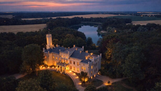 A large, castle-like building with a tower, surrounded by trees and a lake.
