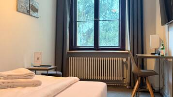 Een hotelkamer met een bed, een bureau met stoel, een radiator, een raam met gordijnen, en een televisie.