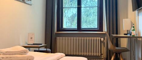 Habitación de hotel con cama, escritorio con silla, radiador, ventana con cortinas y televisor.