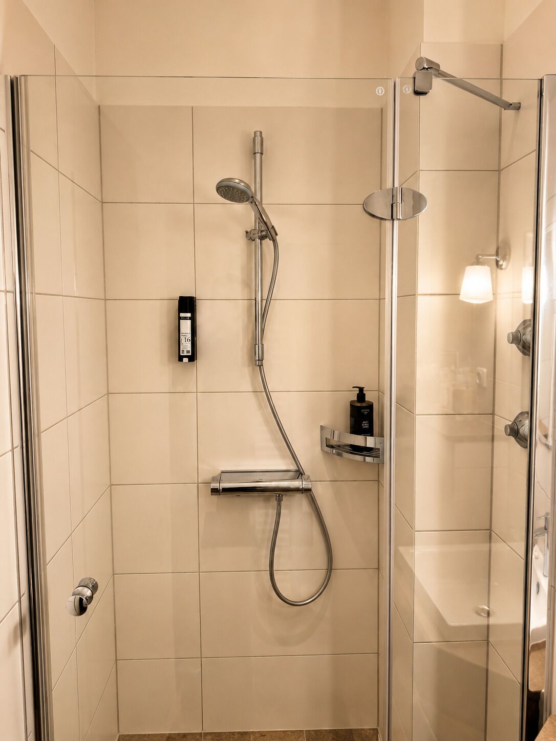 Ducha con cerramiento de vidrio, cabezal de ducha de mano, grifo empotrado y dispensador de jabón.
