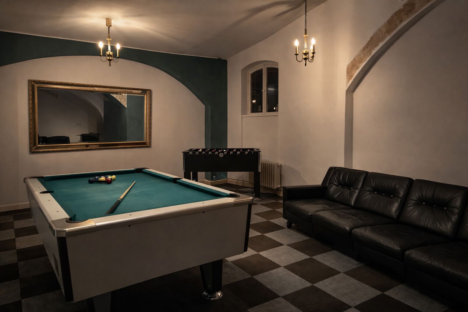 Een pooltafel met een tafelvoetbal en een leren bank in een ruimte met geruit vloer en een spiegel aan de muur.
