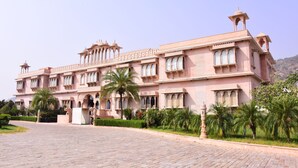 A pink building with arched windows and a central dome, surrounded by palm trees and a paved courtyard. - Bhanwar Singh Palace (Pushkar)