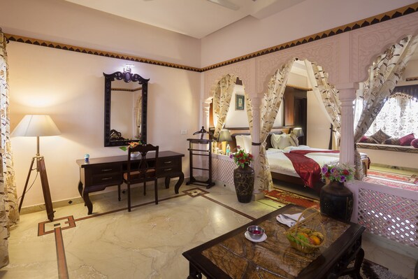 A room with a bed, a desk, a chair, a lamp, a mirror, and a vase with flowers. - Bhanwar Singh Palace (Pushkar)