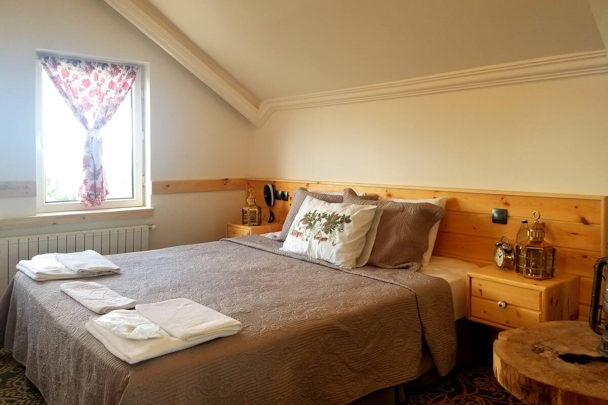 Ein Schlafzimmer mit einem Holzkopfende, einem großen Bett mit einem gemusterten Überwurf, zwei Nachttischen mit Lampen, einem Fenster mit floralen Vorhängen und einem kleinen runden Tischchen mit einer Uhr.