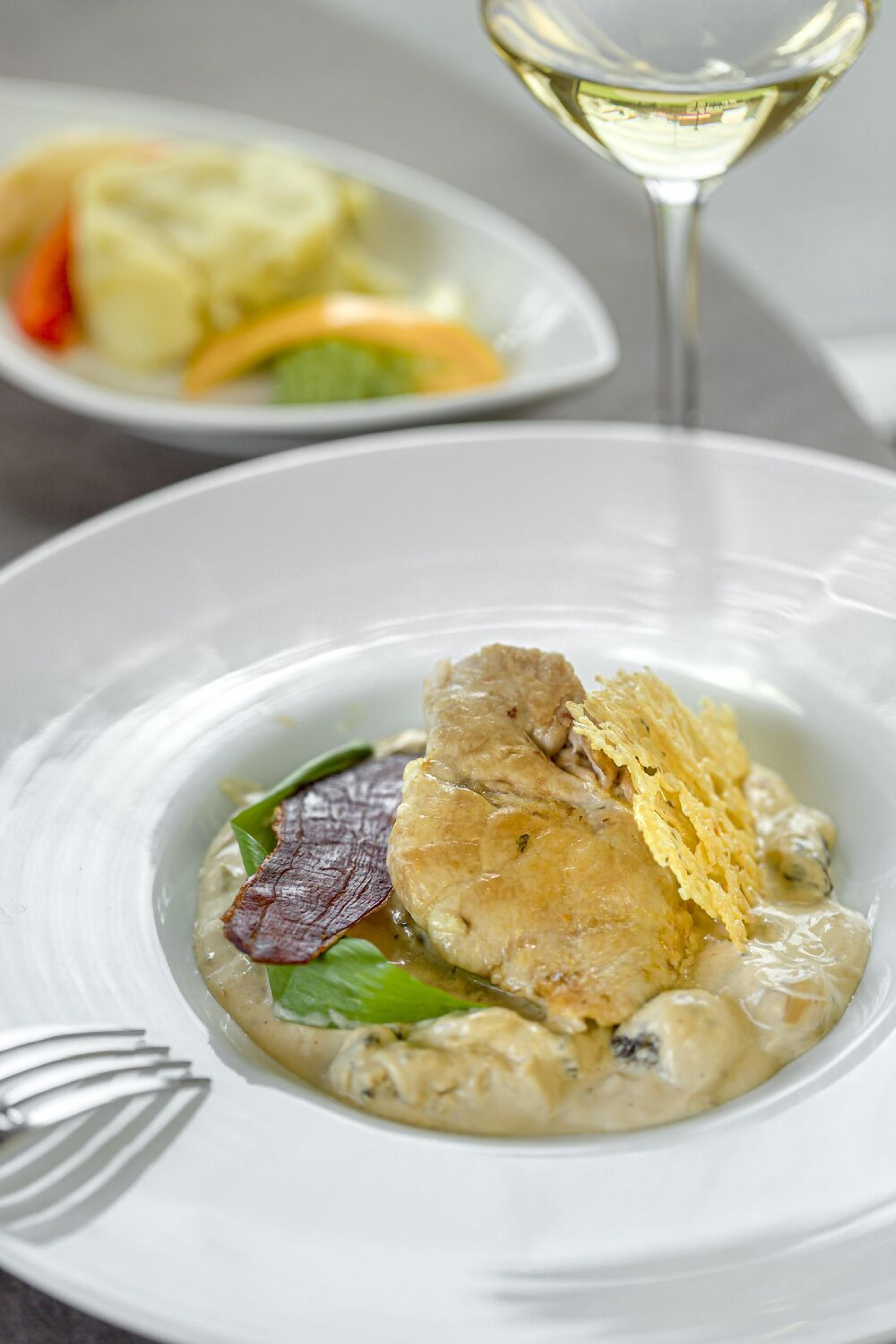 Un plato blanco con un guiso cremoso, una copa de vino blanco y un tenedor.