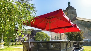 An outdoor seating area with wicker chairs and a red umbrella, overlooking a historic building with a tower. - Hôtel Le Vouglans (Orgelet)