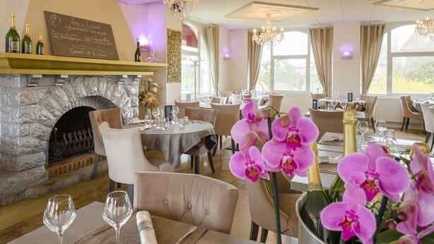 A restaurant with a stone fireplace, tables set for dining, and a chandelier. - Hôtel Le Vouglans (Orgelet)