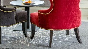 A seating area with a grey tufted sofa and a red velvet chair with gold detailing. A small round table with a glass of beer and a plate of food is in front. - Hôtel Le Vouglans (Orgelet)
