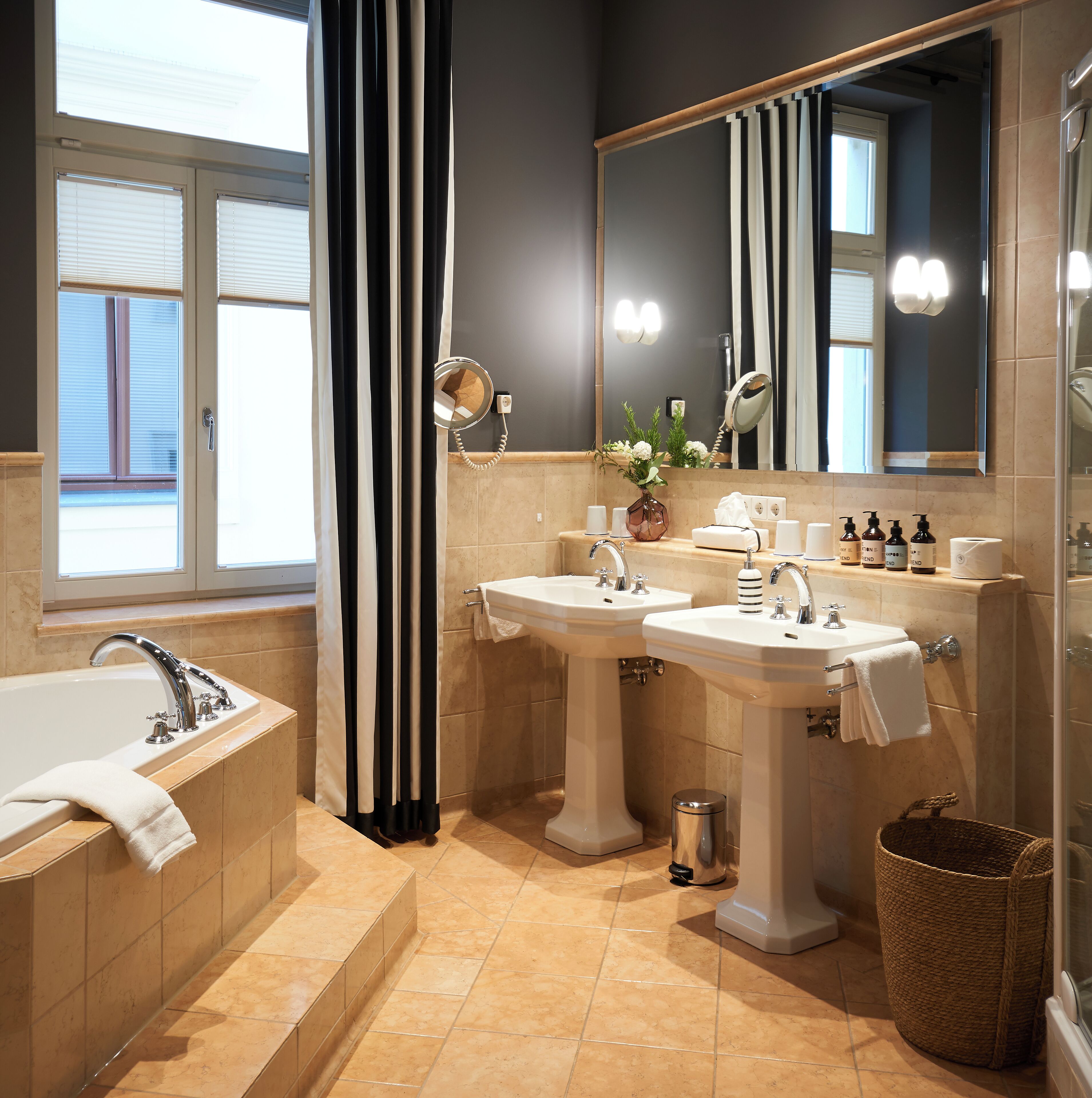 A modern bathroom with a bathtub, two sinks, a large mirror, and a window with blinds.