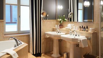 A modern bathroom with a bathtub, two sinks, a large mirror, and a window with blinds.