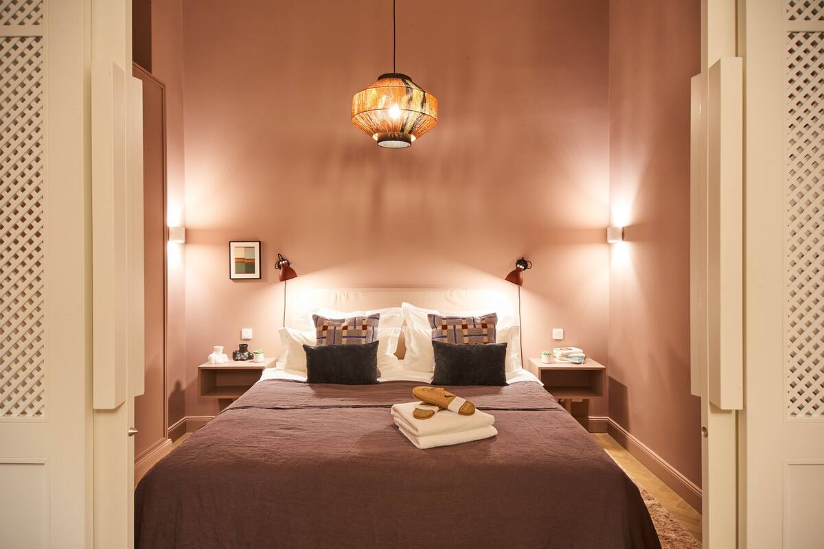 A bedroom with a large bed, two bedside tables, a hanging light fixture, and a wall-mounted shelf.