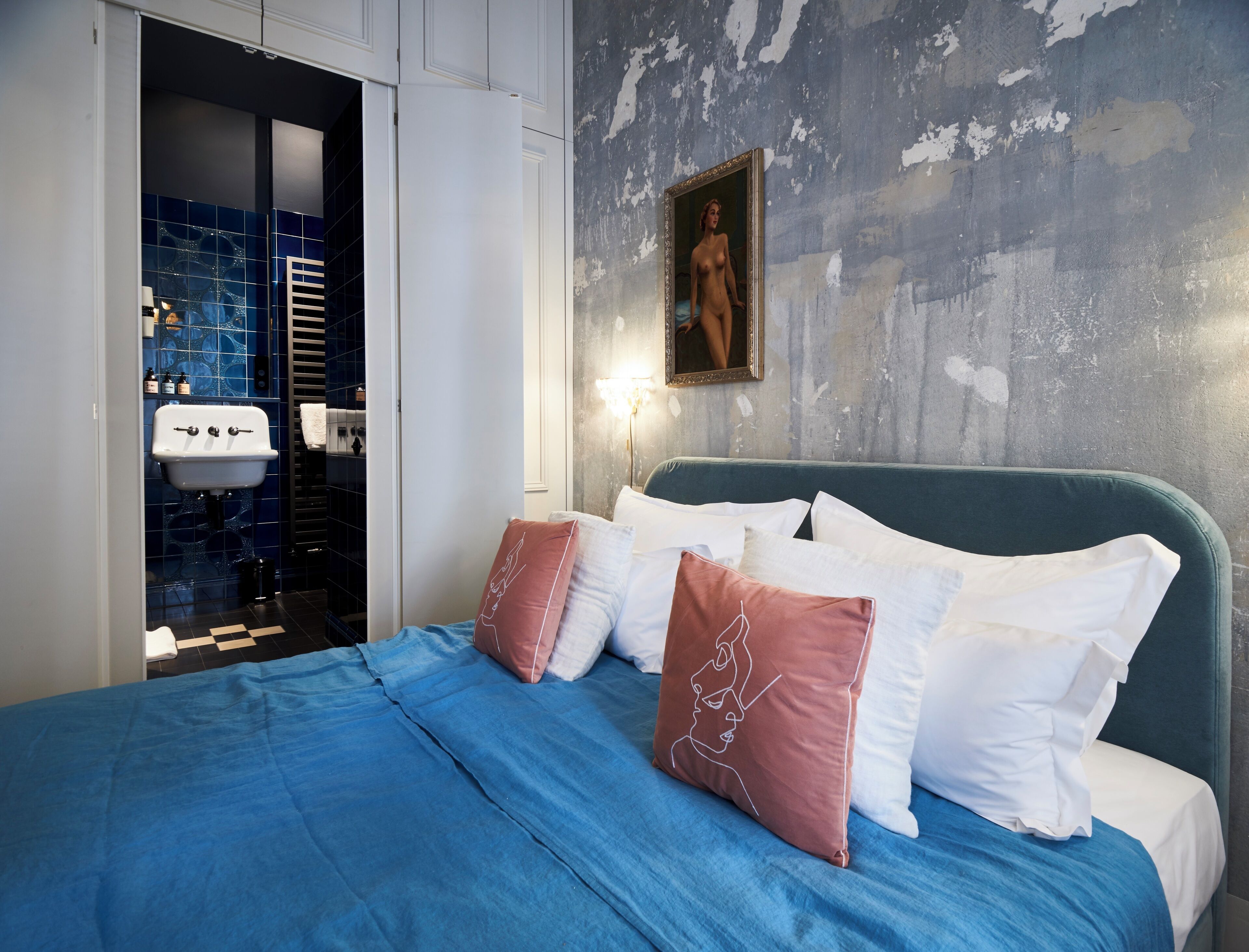 Een slaapkamer met een bed, kussens, een nachtkastje, een schilderij en een badkamer met een wastafel en tegels.