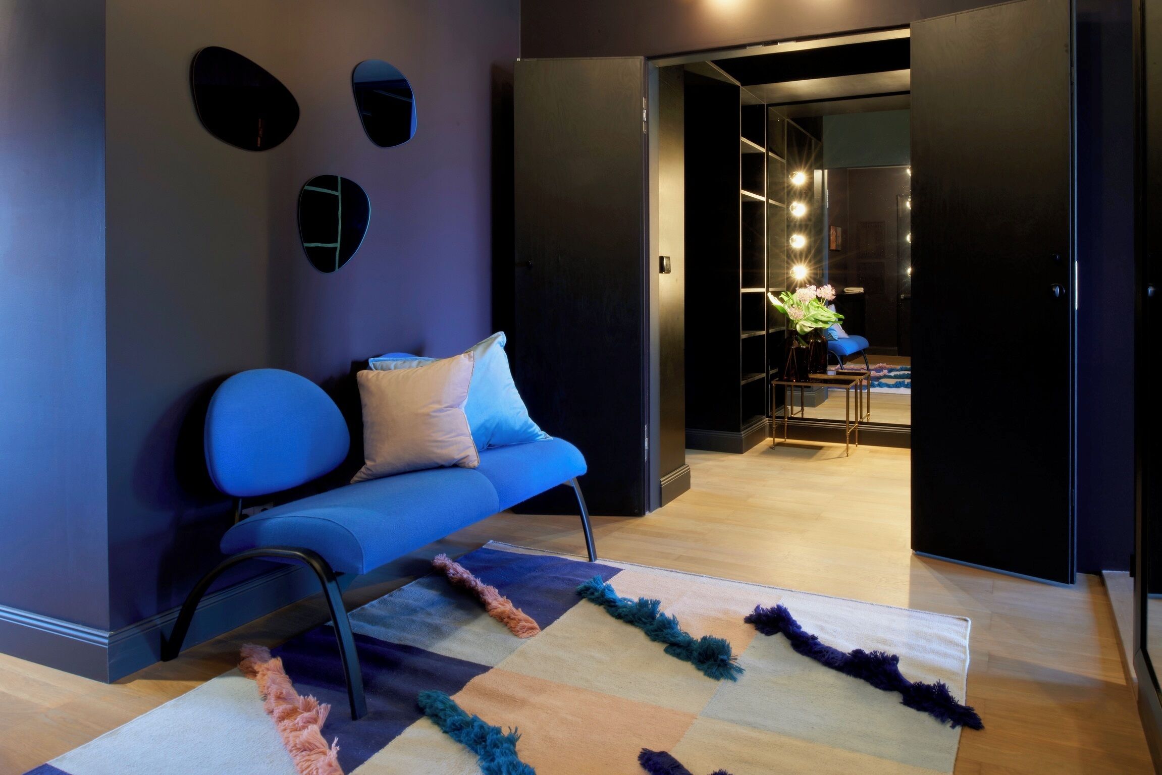 Ein modernes Wohnzimmer mit einem blauen Sofa, einem gemusterten Teppich und einer dunklen Wand mit drei runden Spiegeln.