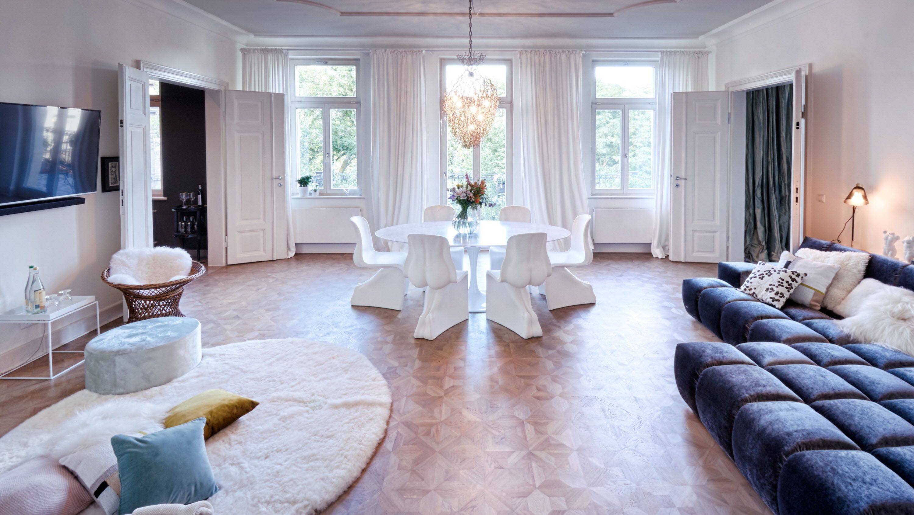 A spacious living room with a large TV, a dining area with a white table and chairs, a blue sofa, and a chandelier.