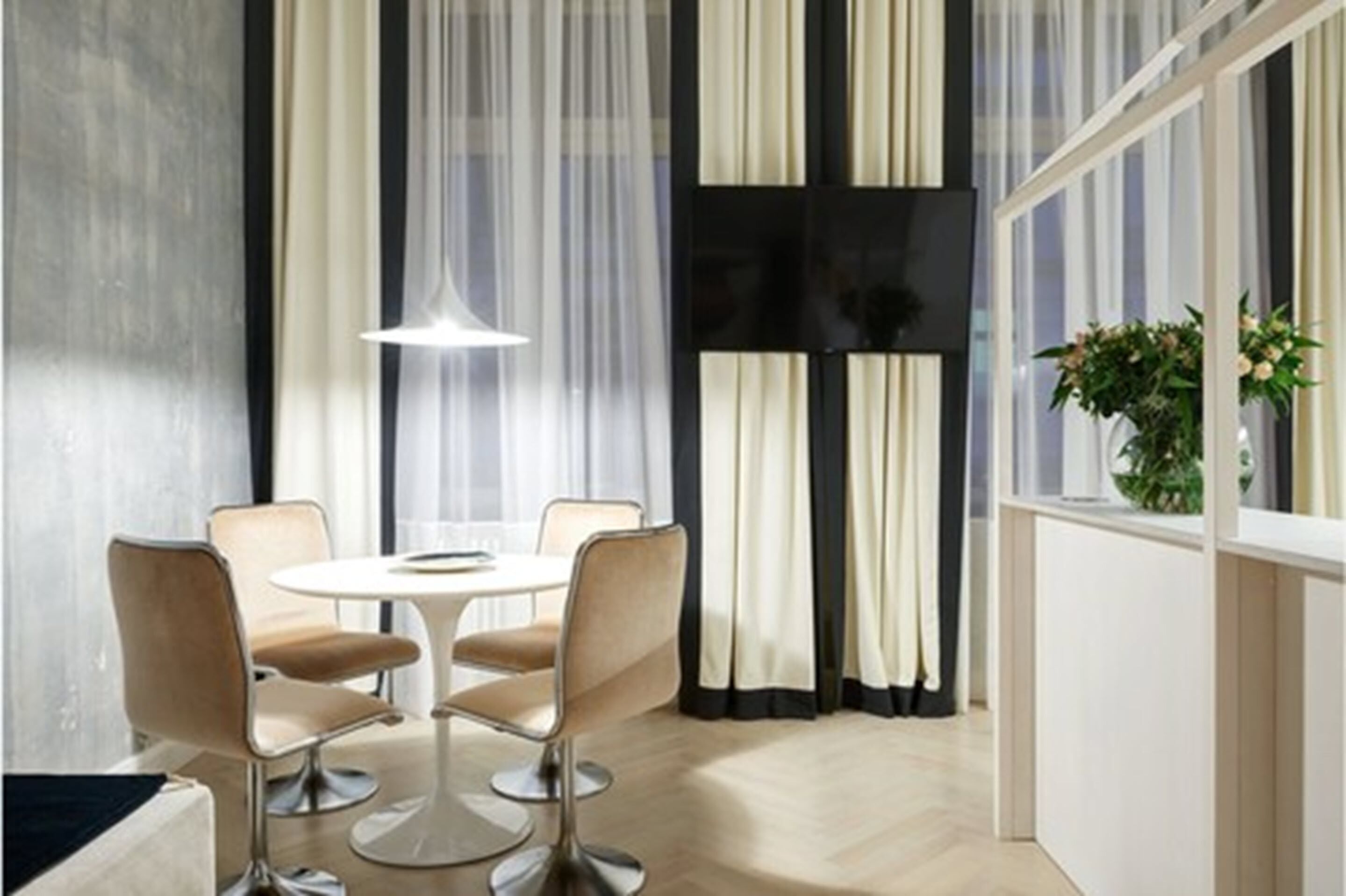 A modern dining area with a round table and four chairs, a large window with sheer curtains, and a potted plant on the windowsill.
