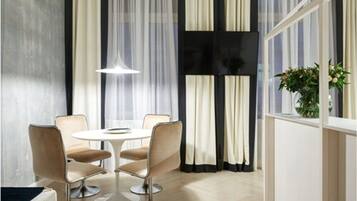 A modern dining area with a round table and four chairs, a large window with sheer curtains, and a potted plant on the windowsill.