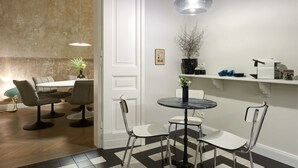 Et moderne spiseområde med et rundt bord, fire stole, en hængende lampe og en vægmonteret hylde med diverse genstande.