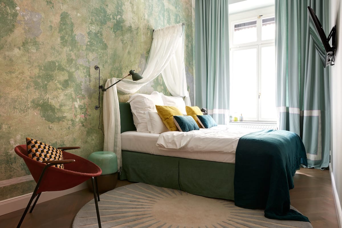 A bedroom with a bed, a chair, a bedside table, a window with curtains, and a wall with a textured finish.