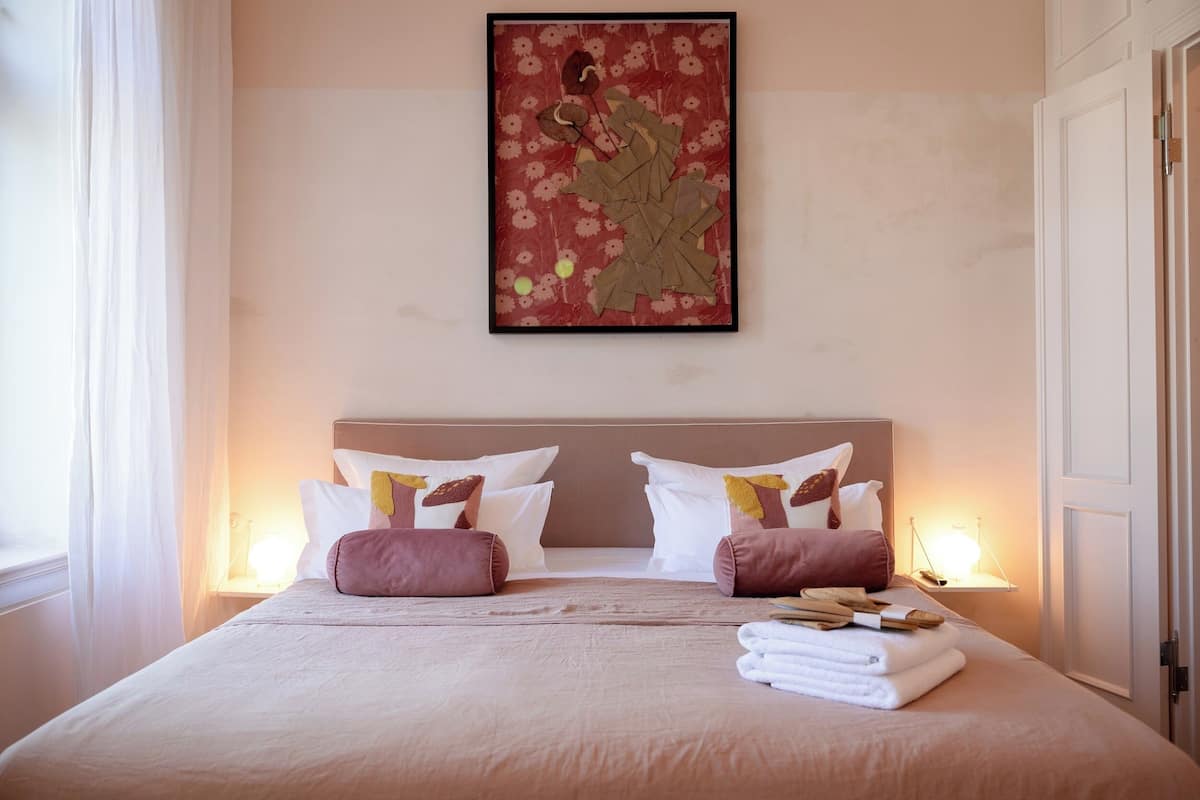 A neatly made bed with white and pink bedding, a framed artwork above, and a bedside table with a lamp.