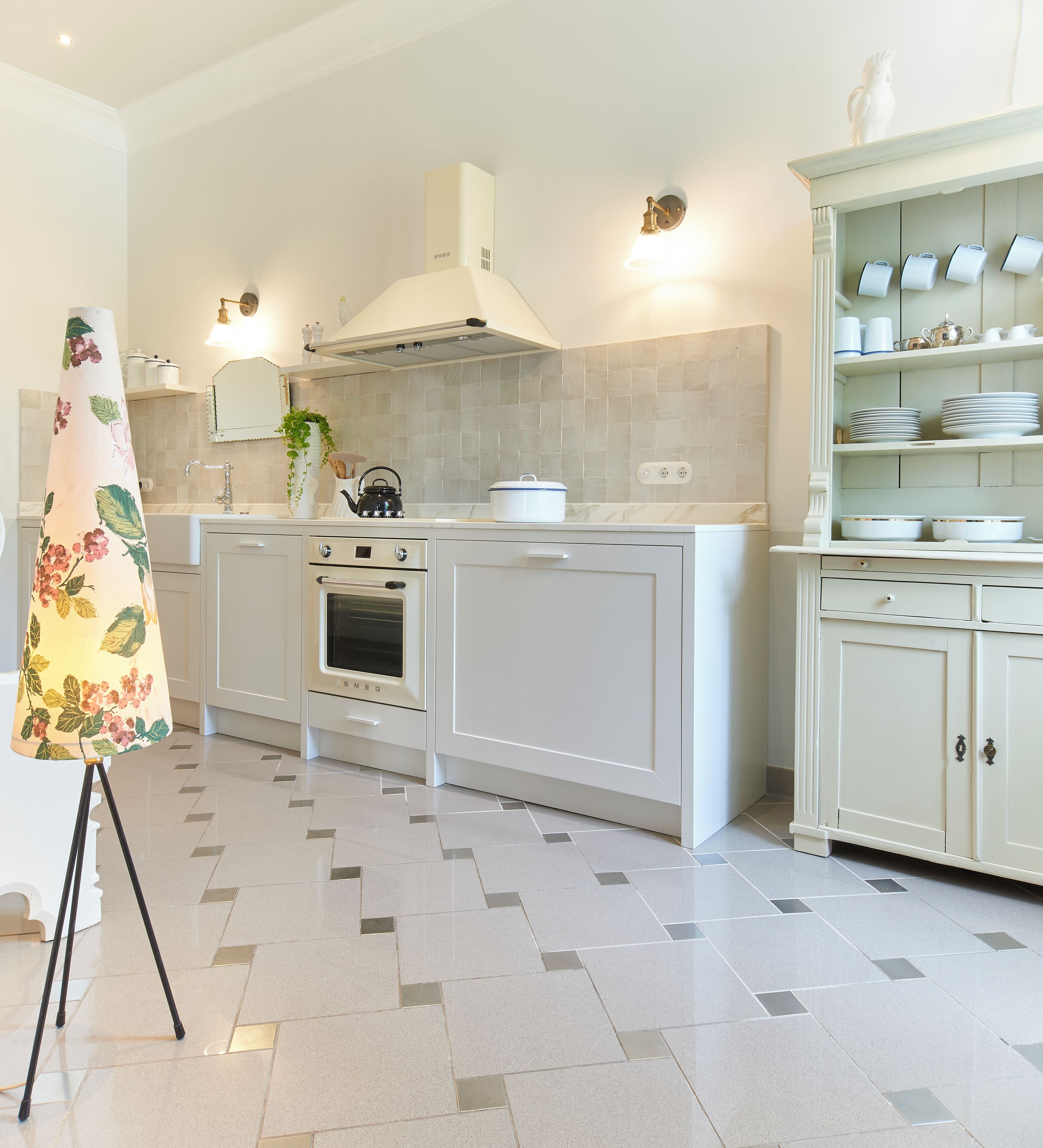 Une cuisine moderne avec des meubles blancs, un four encastré et une cuisinière. On y trouve une lampe décorative et une étagère avec de la vaisselle.