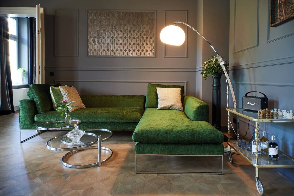 A green sofa with a glass coffee table, a floor lamp, and a bar cart with bottles.