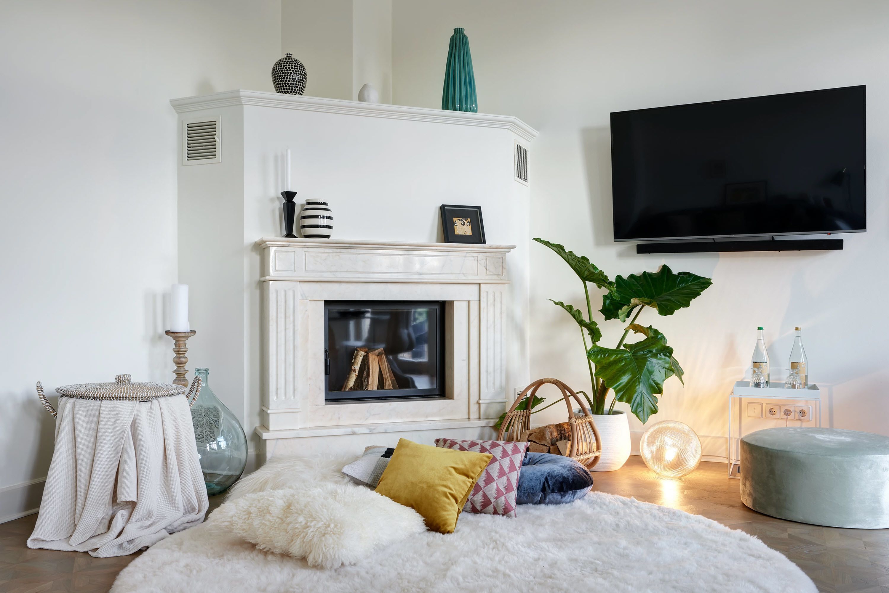 A living room with a fireplace, a television mounted on the wall, a round ottoman, and a large potted plant.