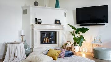A living room with a fireplace, a television mounted on the wall, a round ottoman, and a large potted plant.