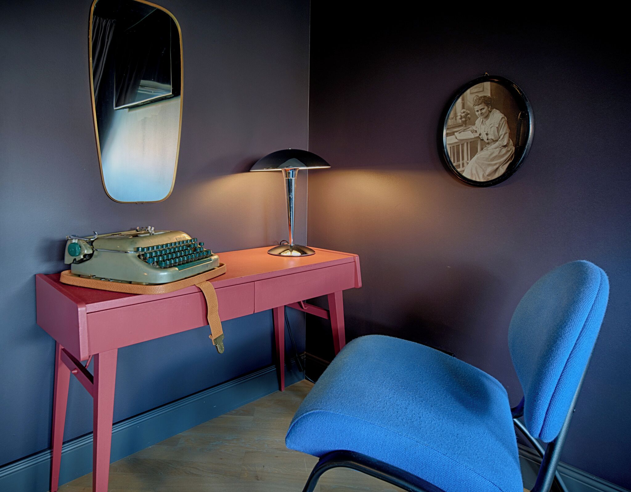 A room with a desk, a typewriter, a lamp, a chair, a mirror, and a framed picture on the wall.
