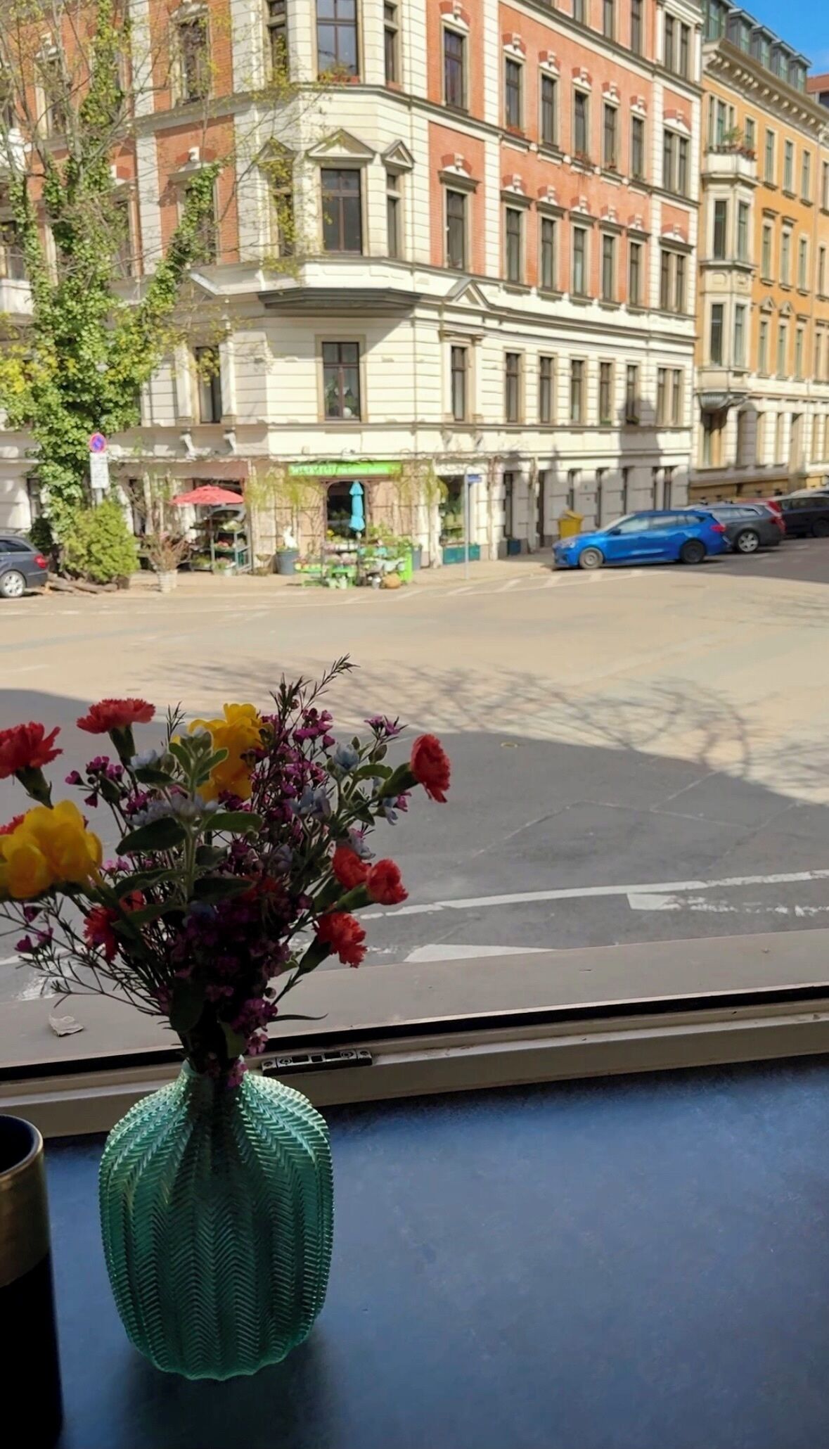 Une vue par la fenêtre sur une rue, avec un vase vert rempli de fleurs posé sur une table.