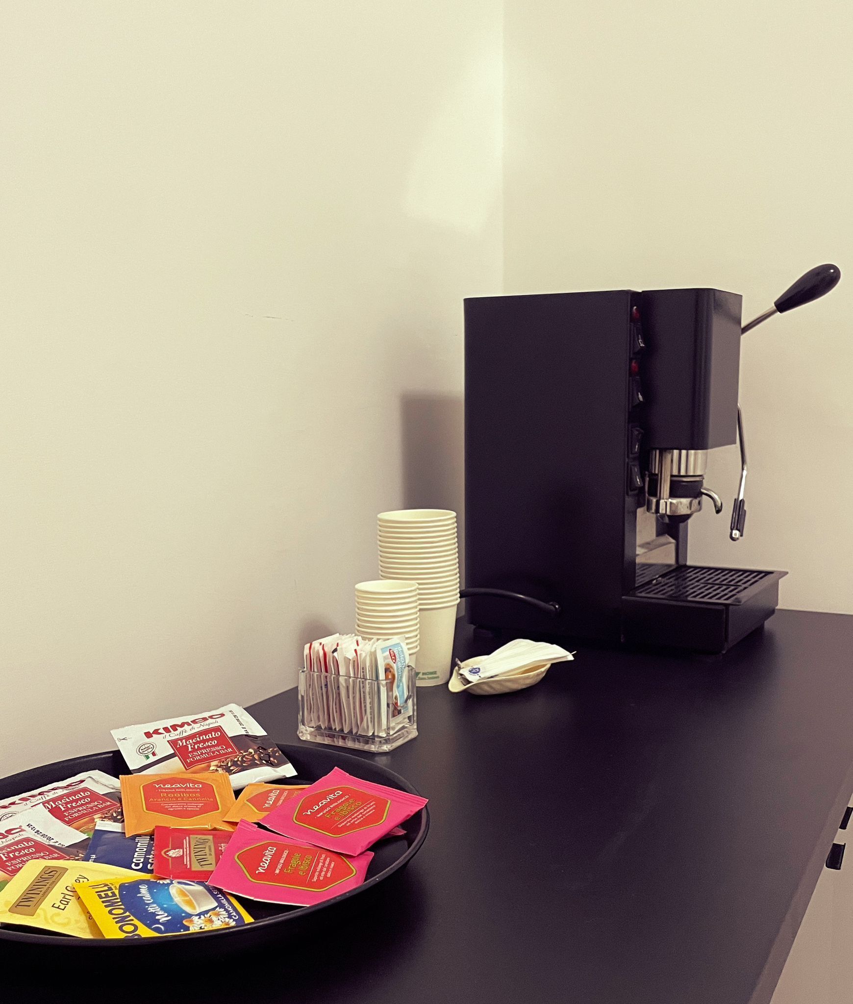 A coffee machine, stacks of white cups, and a tray with coffee packets on a black table.