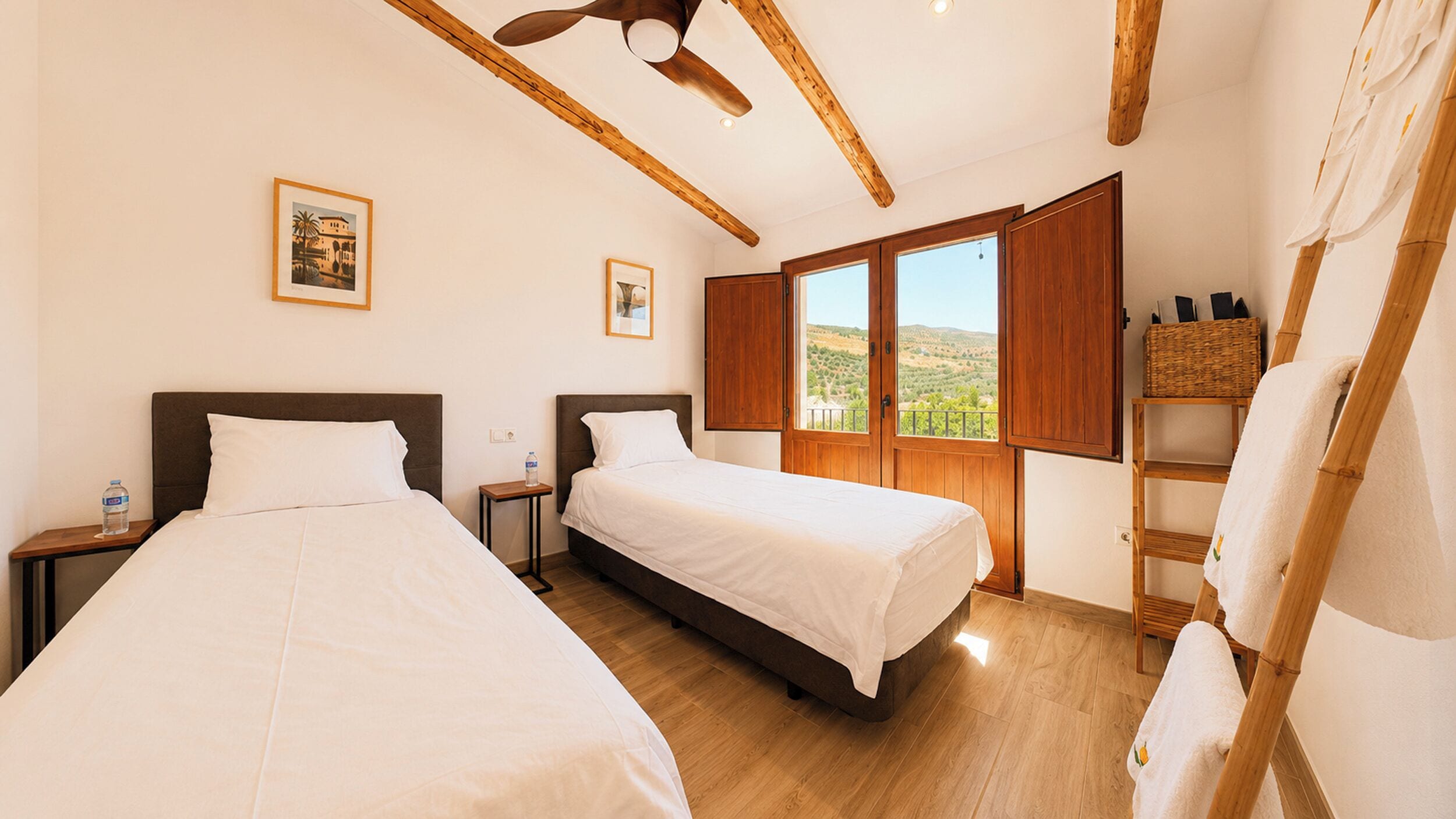 Habitación de hotel con dos camas, vigas de madera, un ventanal con vistas y una pequeña mesa con una botella de agua.
