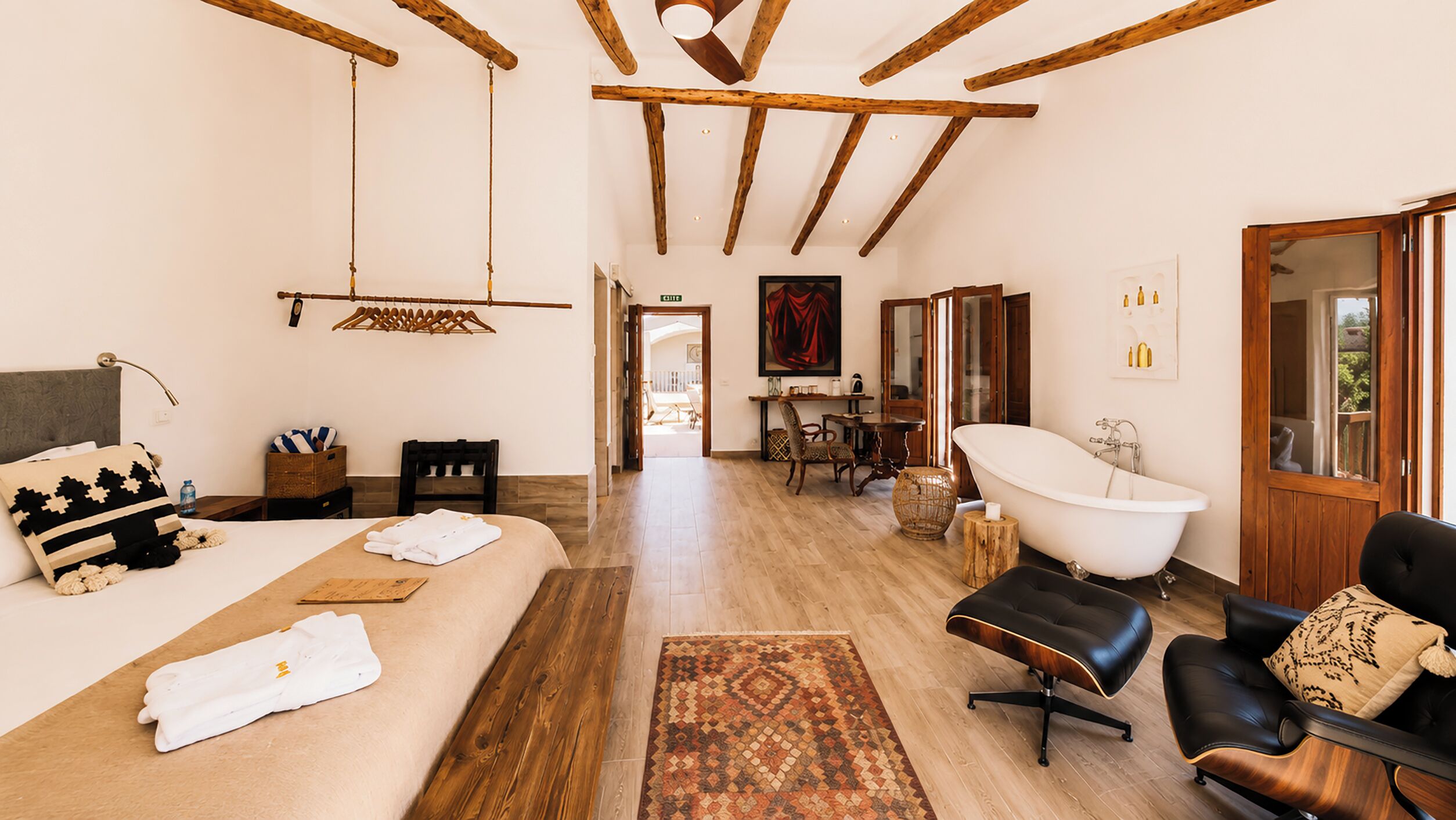 A spacious room with a large bed, a sitting area with a black leather sofa, a white bathtub, and a wooden ceiling.