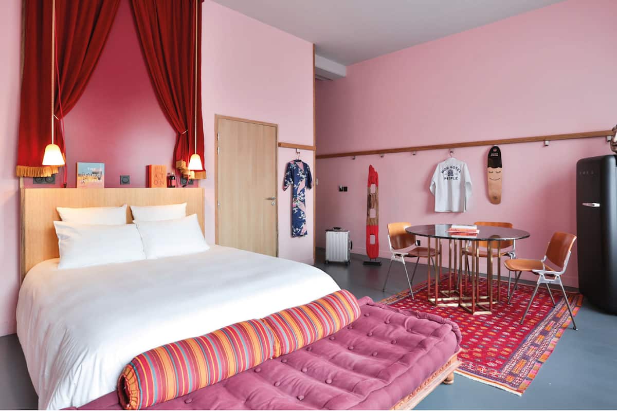 Habitación de hotel con una cama grande, una mesita con sillas, una cortina roja y un perchero en la pared.