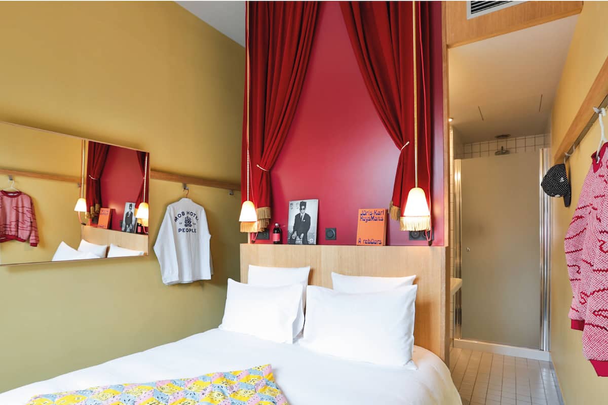 Habitación de hotel con cama, espejo, mesita de noche con lámpara, cortina roja y zona de ducha.