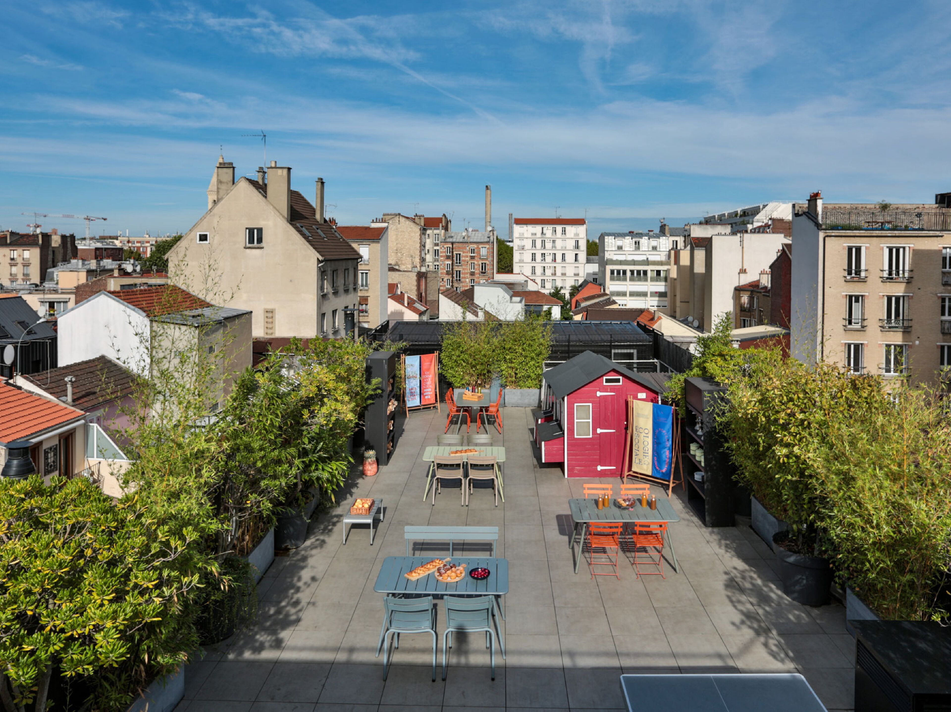 Une terrasse extérieure avec des tables et des chaises, entourée de verdure et avec des bâtiments en arrière-plan.