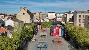 Une terrasse extérieure avec des tables et des chaises, entourée de verdure et avec des bâtiments en arrière-plan.