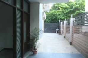 A narrow corridor with a potted plant, a gated entrance, and a person sitting on a bench. - Mint City Center Suites (Noida)