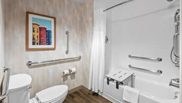 A bathroom with a toilet, a shower with a glass door, a handrail, a towel rack, and a framed picture on the wall.