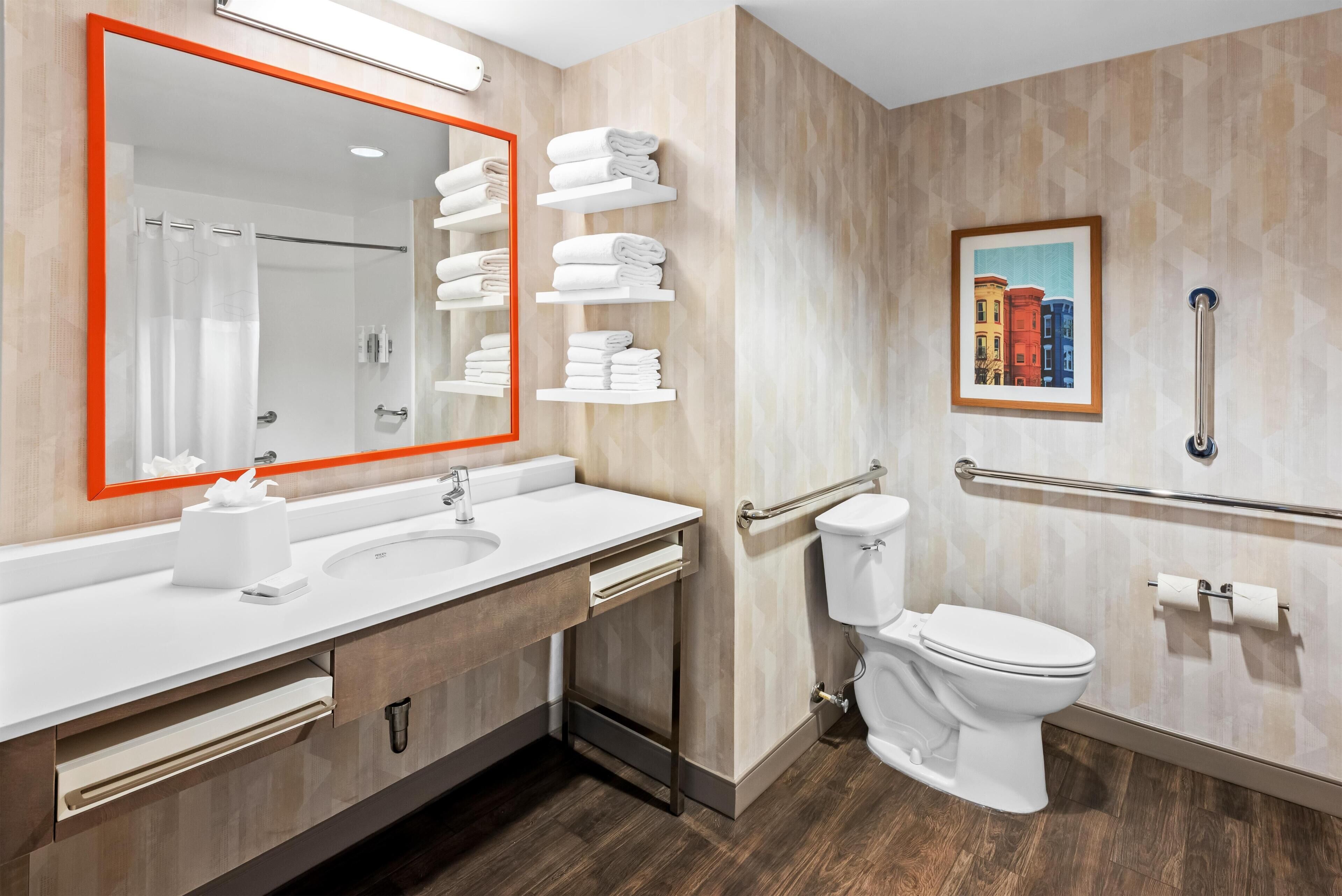 Un baño moderno con un espejo grande, lavamanos blanco y un inodoro. Hay toallas en un estante, un pasamanos y un cuadro en la pared.