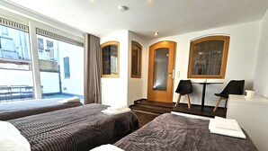 Hotellihuone, jossa on kaksi sänkyä, ovi, jossa on himmeä lasipaneeli, pieni pöytä ja kaksi tuolia sekä näkymä ulkopuolella olevalle parvekkeelle.