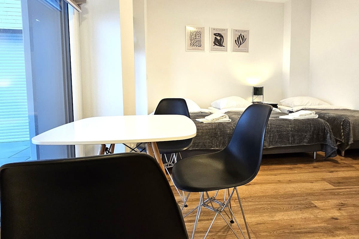 Une chambre d’hôtel avec un plancher en bois, une table blanche, deux chaises noires, un lit recouvert d’une couverture grise et trois œuvres d’art encadrées accrochées au mur.